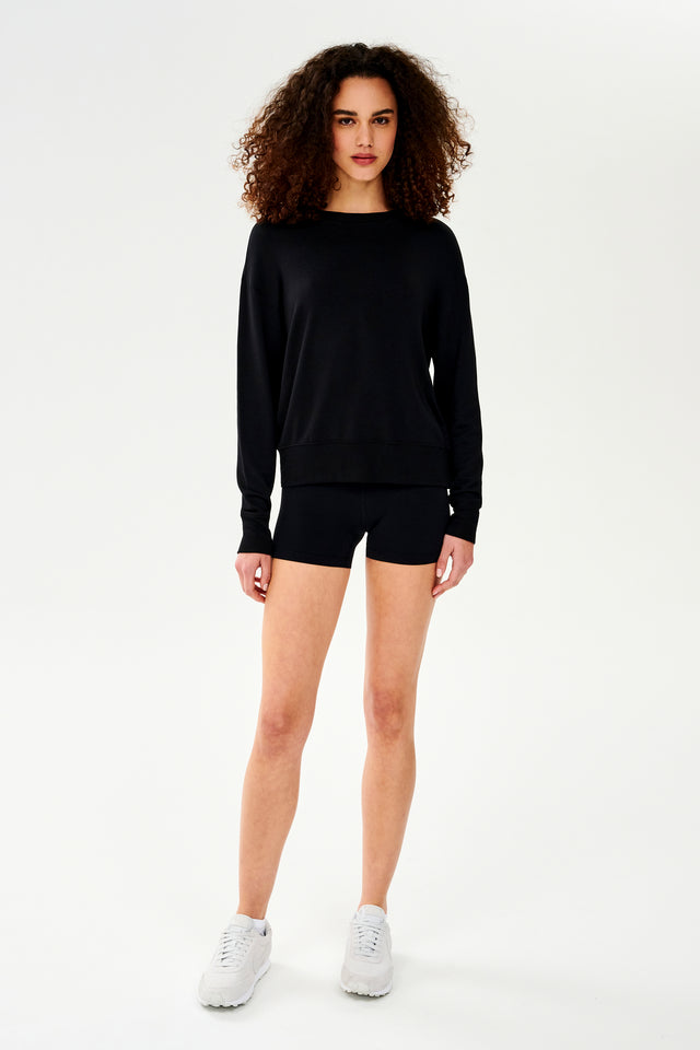 Full front view of woman with curly dark brown hair wearing a black crewneck sweatshirt and black shorts paired with cream shoes