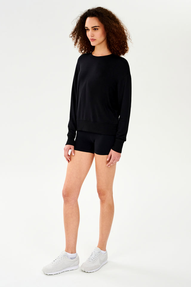Full front side view of woman with curly dark brown hair wearing a black crewneck sweatshirt and black shorts paired with cream shoes