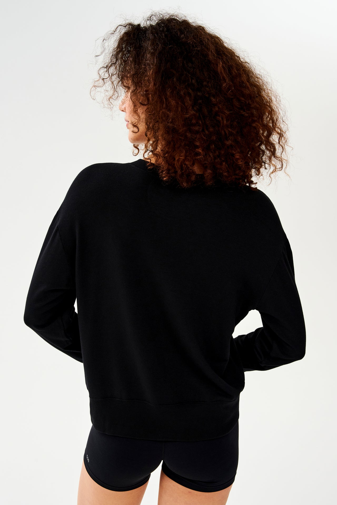 Back view of woman with curly dark brown hair wearing a black crewneck sweatshirt and black shorts