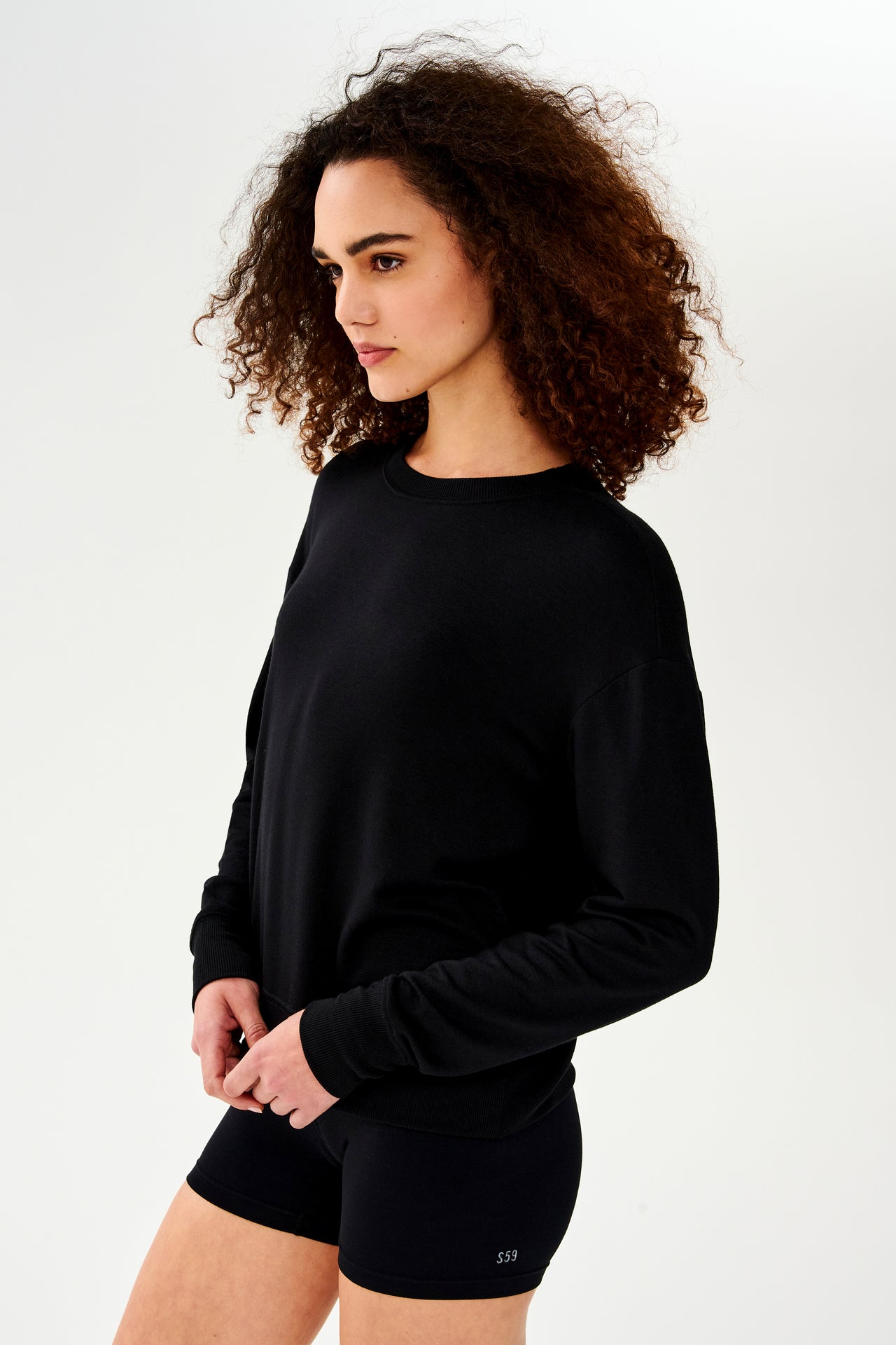 Front side view of woman with curly dark brown hair wearing a black crewneck sweatshirt and black shorts 