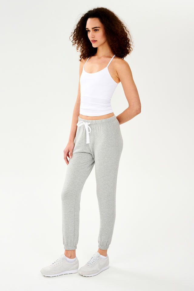 Full front side view of woman with dark curly hair wearing light grey jogger with white drawstring and white cropped tank top paired with creme shoes