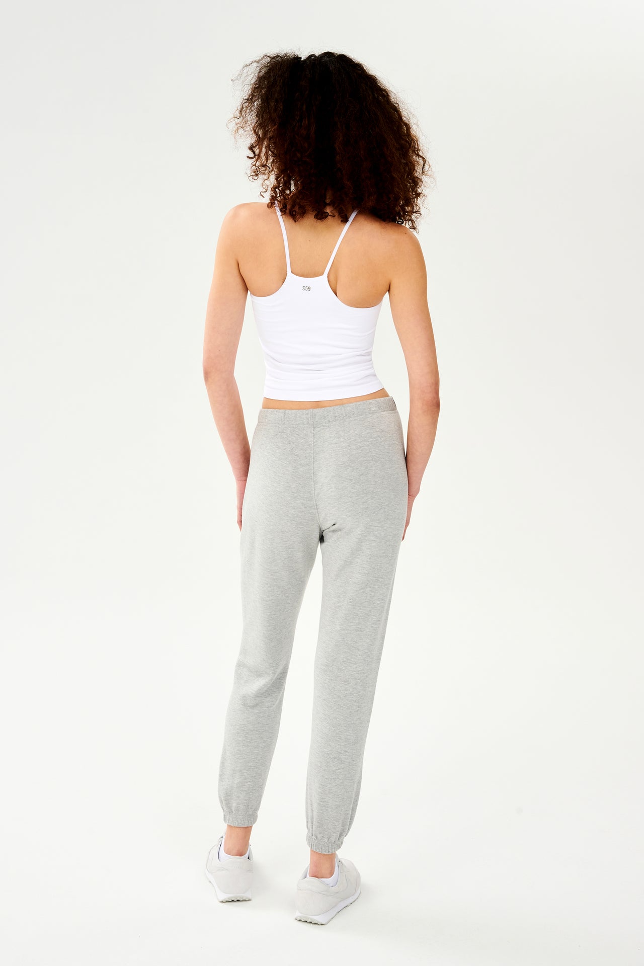 Full front view of woman with dark curly hair wearing light grey jogger with white drawstring and white cropped tank top with racerback paired with creme shoes
