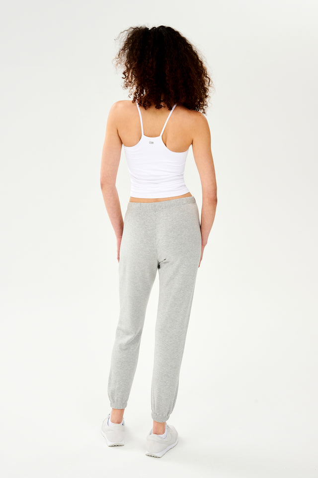 The back view of a woman wearing grey sweatpants and a SPLITS59 Loren Seamless Waist Length Tank in white, with a shelf bra, ready for gym workouts.