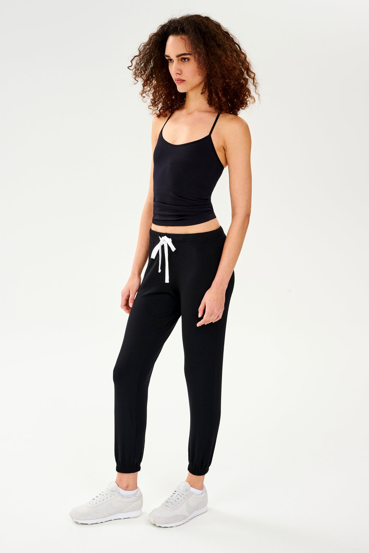 Full front side view of woman with dark curly hair wearing black jogger sweatpant with white drawstring and black cropped tank top paired with white shoes