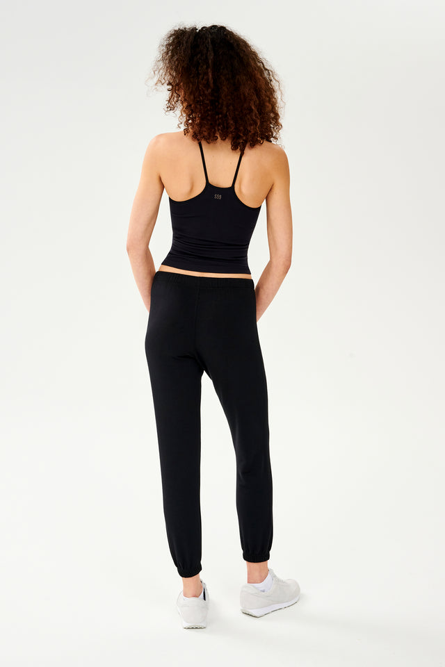 Full back view of woman with dark curly hair wearing black jogger sweatpant with white drawstring and black cropped tank top with racerback paired with white shoes