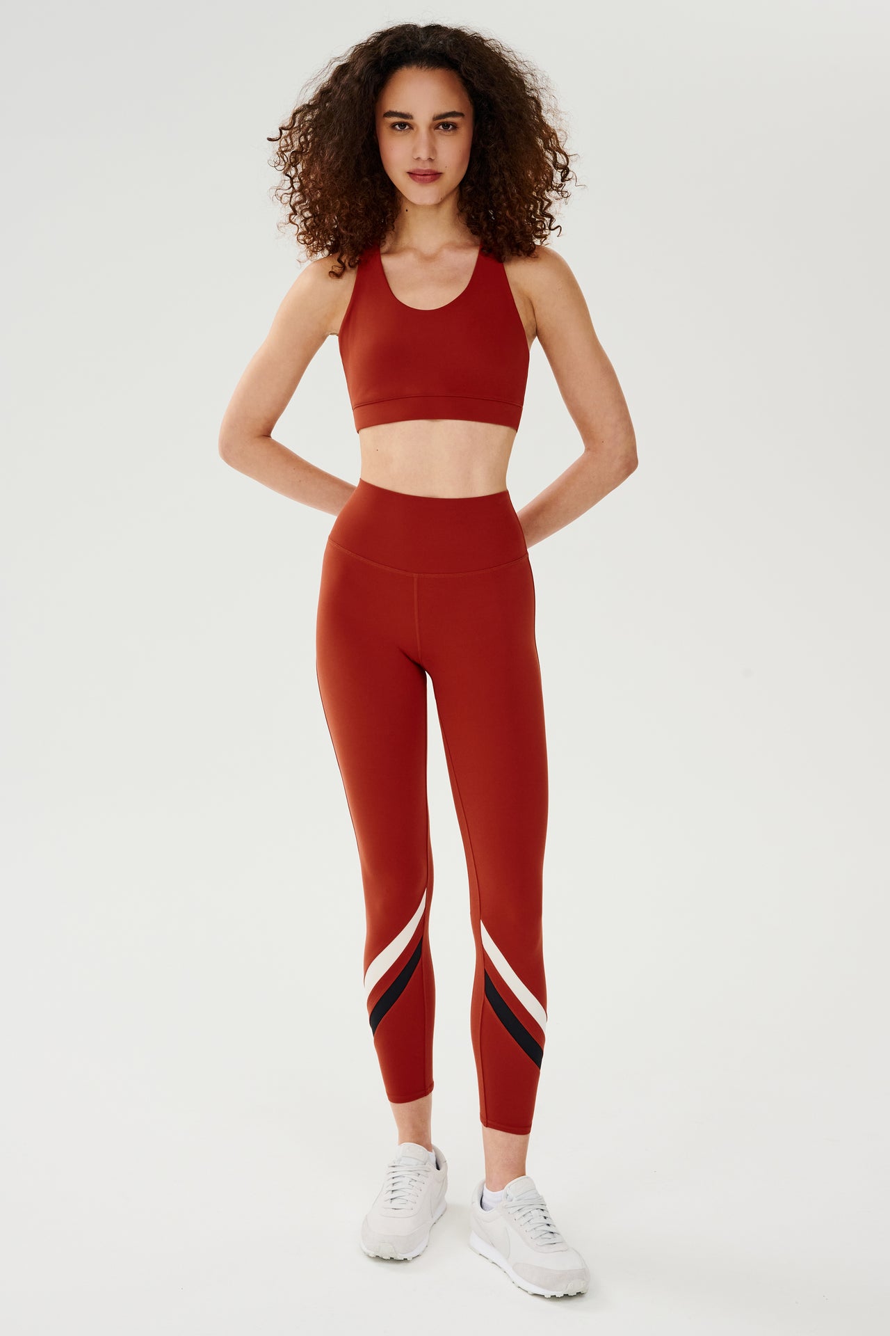 Full front view of girl wearing red thick straps sports bra with red leggings with black and white stripes on shins and white shoes