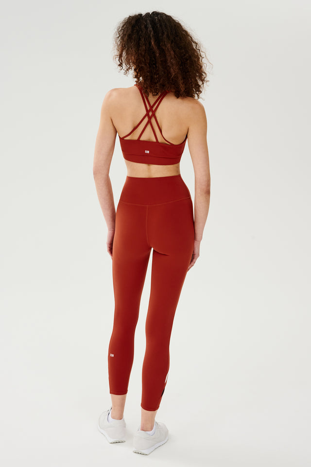 Full back view of girl wearing red thick straps sports bra with red leggings with black and white stripes on shins and white shoes