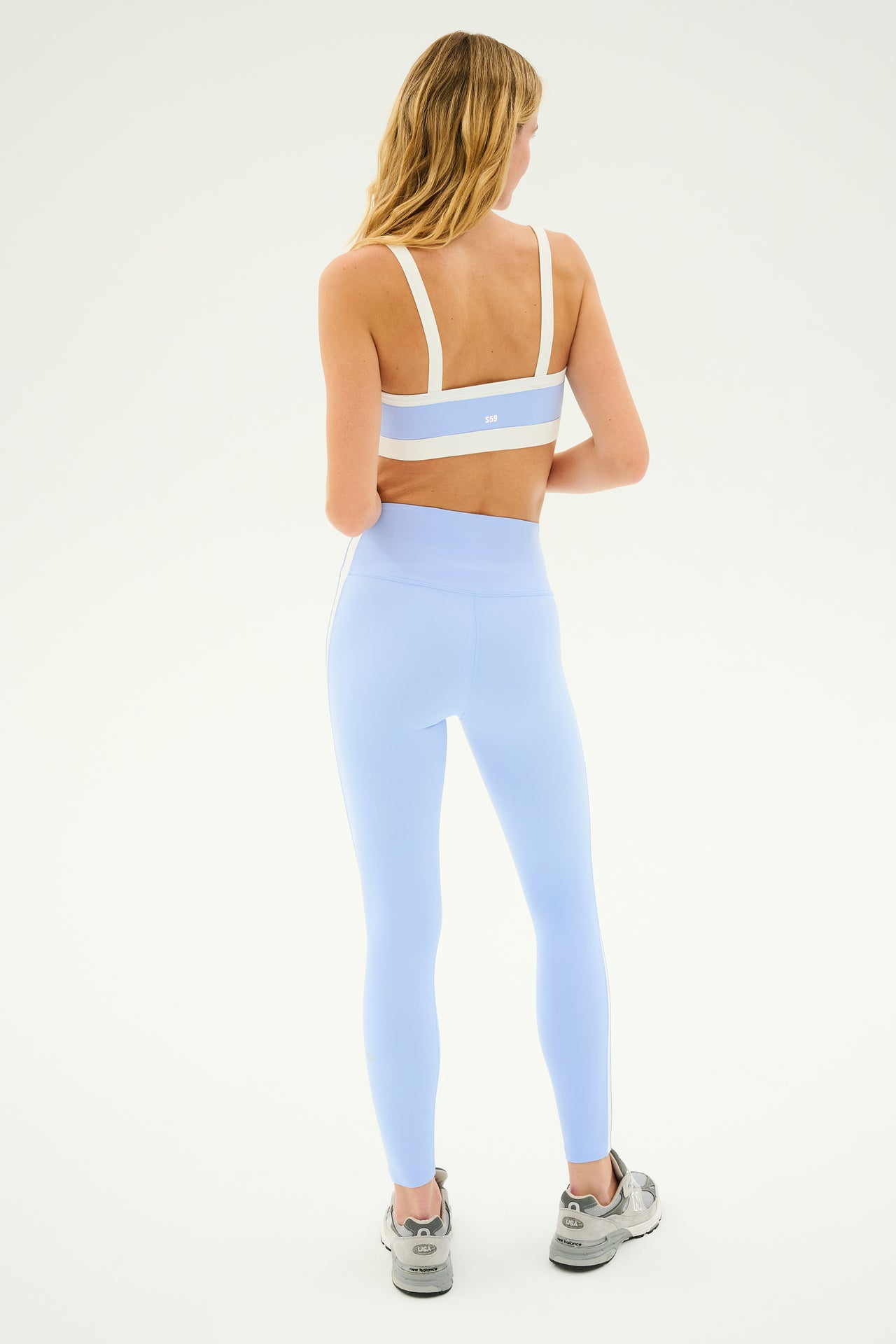 A person wearing the Clare High Waist Rigor 7/8 leggings in Skylight/White from SPLITS59, crafted from a comfortable blend of nylon and spandex, stands facing away to showcase the back of the outfit. They are also wearing grey athletic shoes.