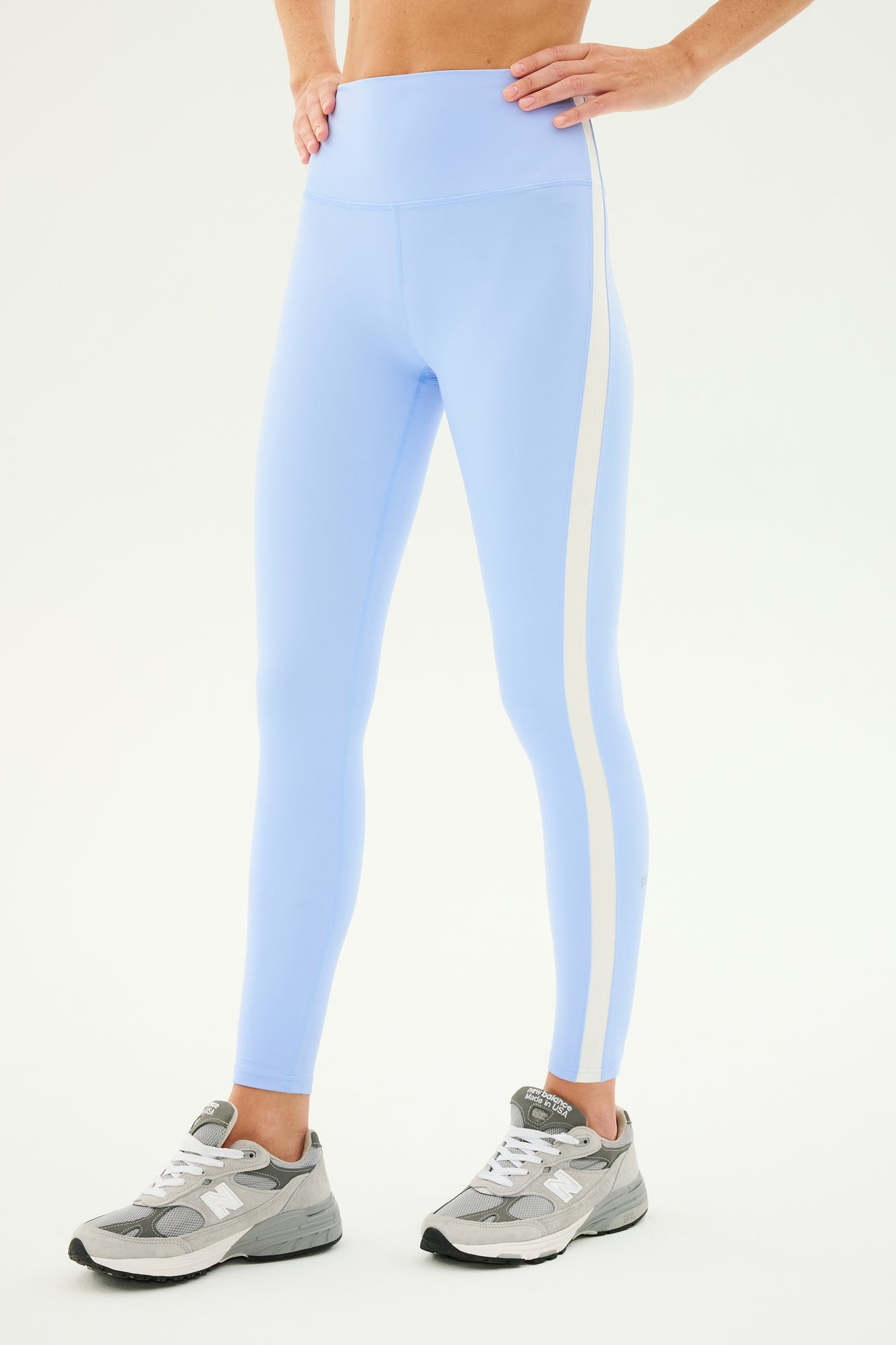 The person is wearing the Clare High Waist Rigor 7/8 leggings in Skylight/White by SPLITS59, complemented by gray athletic shoes. Standing with hands on hips, the individual is depicted from midriff to feet against a plain background.