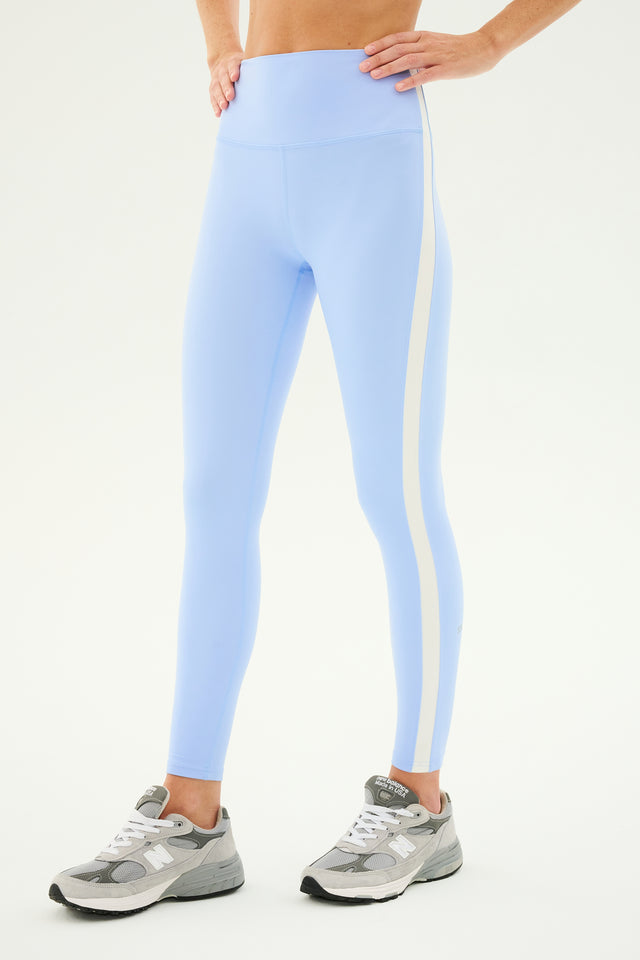 The person is wearing the Clare High Waist Rigor 7/8 leggings in Skylight/White by SPLITS59, complemented by gray athletic shoes. Standing with hands on hips, the individual is depicted from midriff to feet against a plain background.