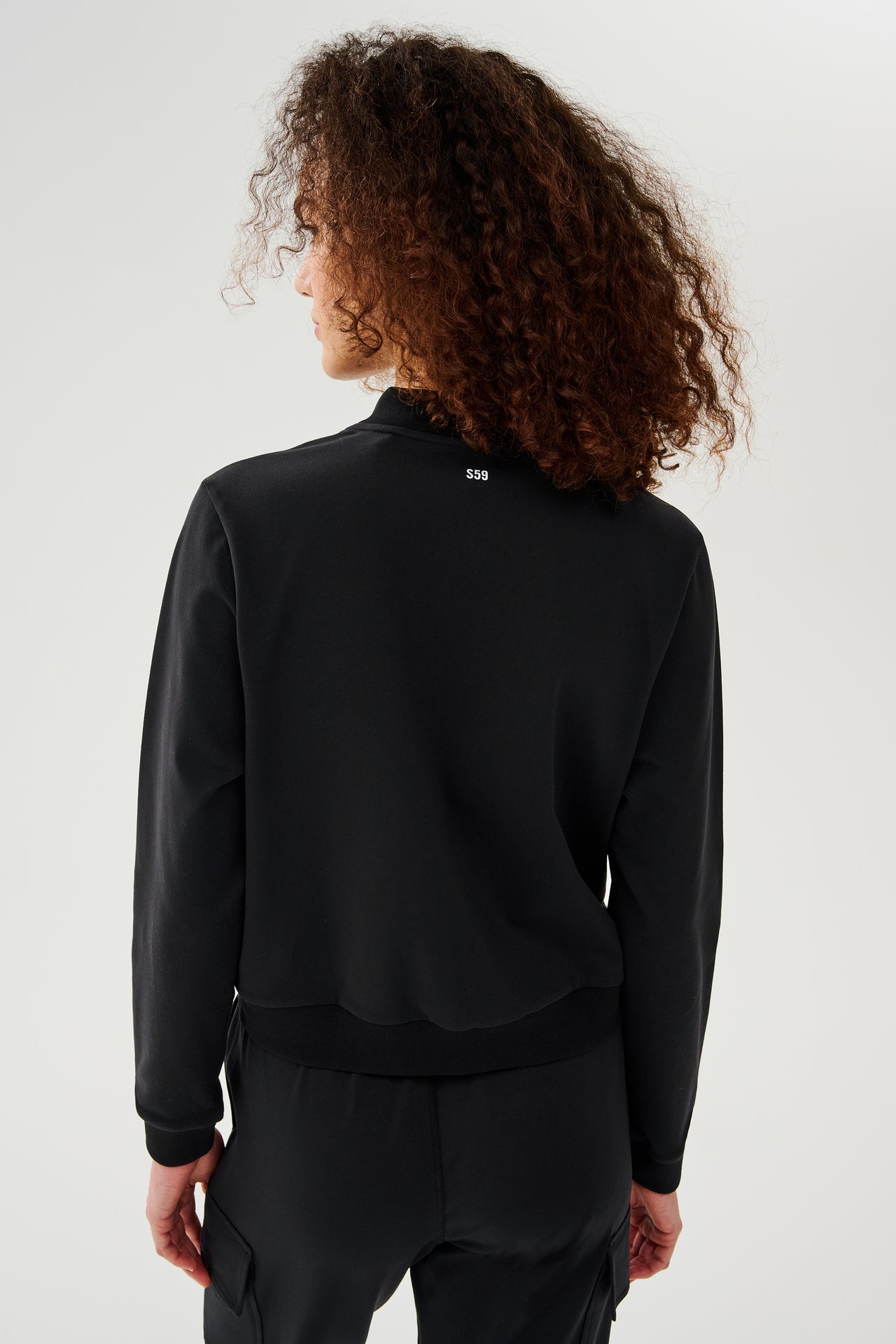 Back view of woman with curly dark brown hair wearing a black jacket zipped open and black bra with black cargo pants 