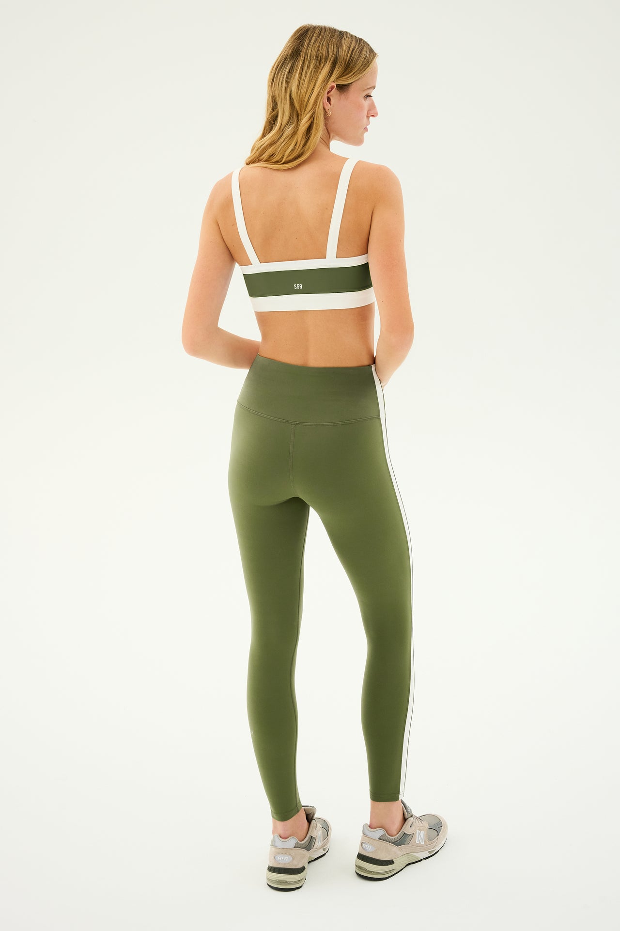 A person with long hair is shown from the back wearing a green sports bra, matching SPLITS59's Clare High Waist Rigor 7/8 leggings in Olive/White, and gray sneakers.