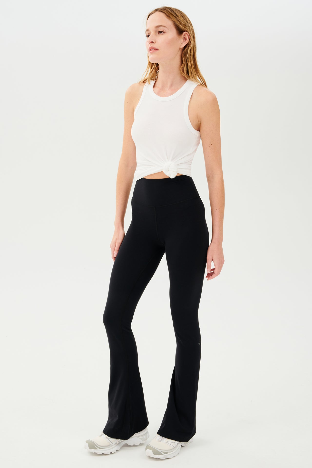 A woman wearing SPLITS59 Raquel High Waist Airweight Flare leggings in black and a white tank top.
