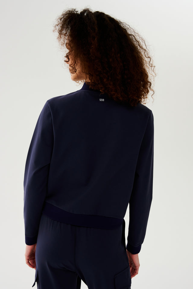 Back view of woman with curly dark brown hair wearing a dark blue jacket with white logo S59 paired with black bra with dark blue cargo pants