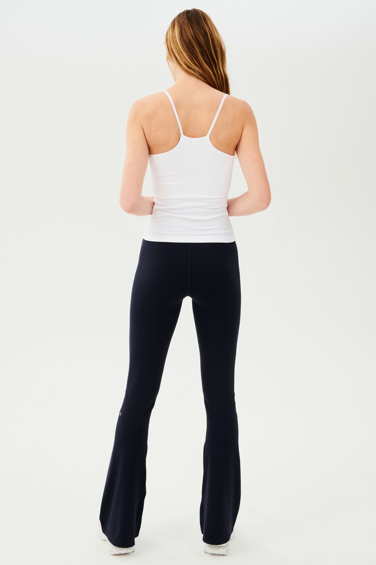 The back view of a woman wearing a white tank top and black leggings made from SPLITS59's Raquel High Waist Airweight Flare - Indigo fabric.