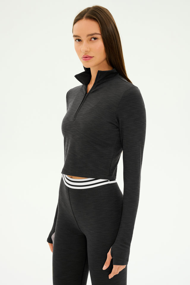 A person with long hair is wearing the SPLITS59 Stevie Active Rib Half Zip in Heather Graphite, paired with black athletic leggings featuring a white waistband. This pullover, made from active rib fabric and designed with a stand-up collar, is perfect for gym workouts.