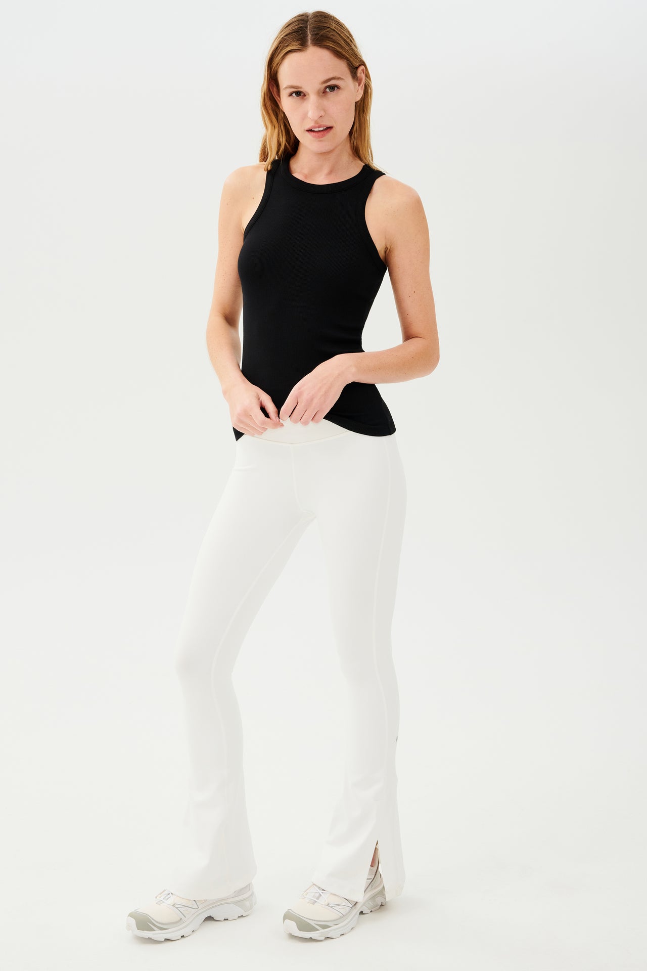 Full front view of woman with blonde straight hair wearing white high waist below ankle length legging with wide flared bottoms with split hem flare opening and black racerback tank top. Paired with white and gray shoes.