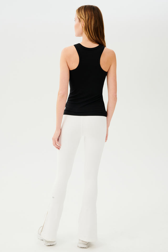 Full back view of woman with blonde straight hair wearing white high waist below ankle length legging with wide flared bottoms with split hem flare opening and black S59 logo on back of left calf and black racerback tank top. Paired with white and gray shoes.