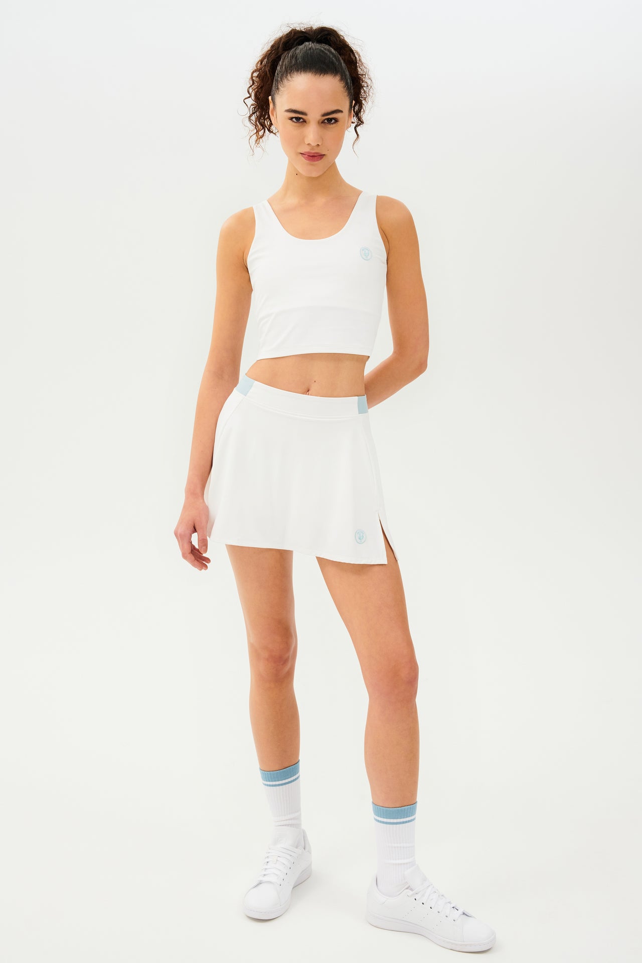 Front full view of woman with dark curly hair in a ponytail wearing a cropped white tank bra with white skort with teal details on the waistband and logo. Model is wearing white socks with teal stripe and all white shoes