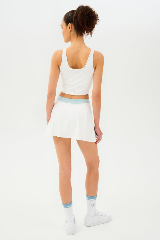Back full view of woman with dark curly hair in a ponytail wearing a cropped white tank bra with white skort with teal details on the waistband and logo.Model is wearing white socks with teal stripe and all white shoes