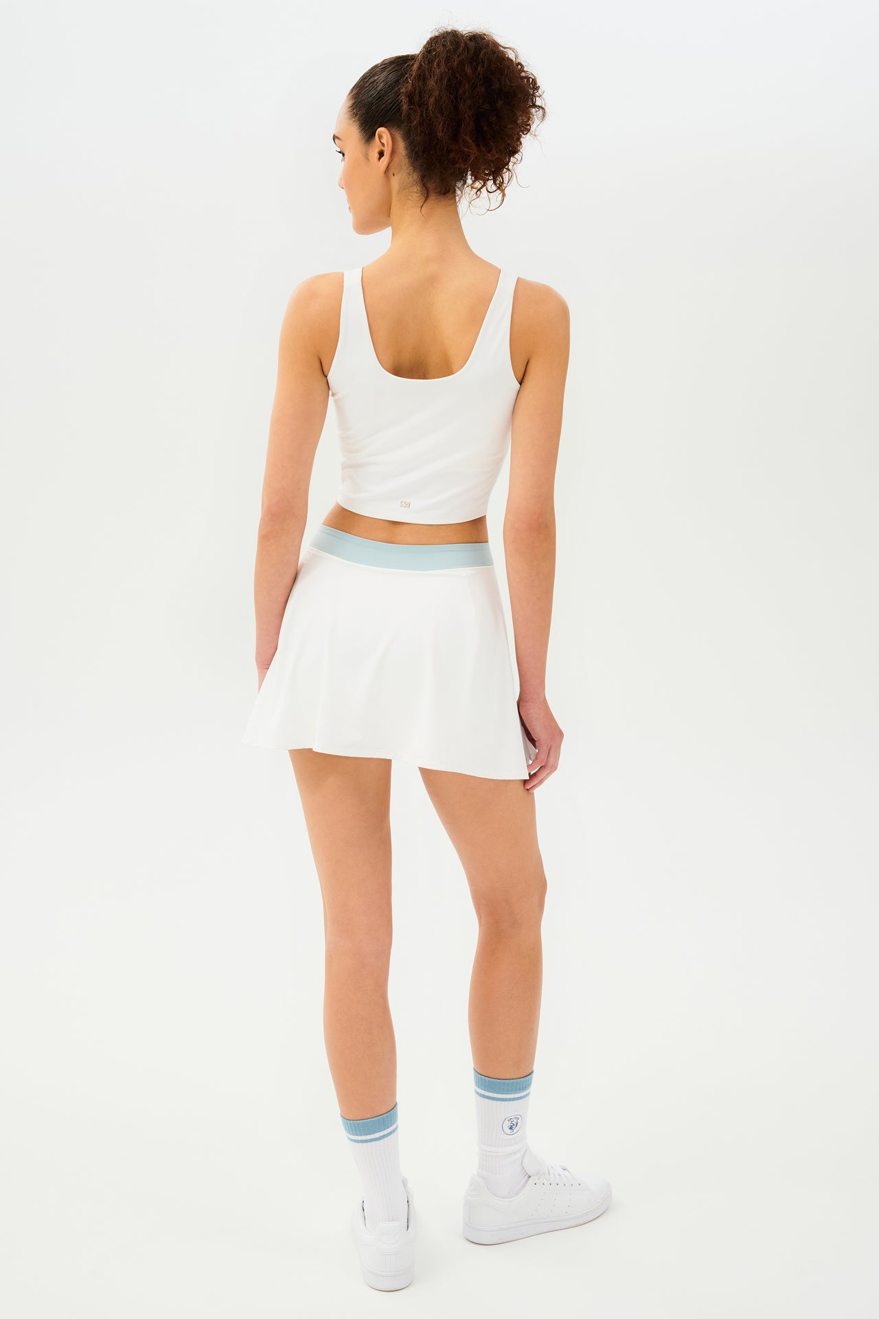 Front back view of woman with dark curly hair in a ponytail wearing a cropped white tank bra with white skort with teal details on the waistband and logo. Model is wearing white socks with teal stripe and all white shoes