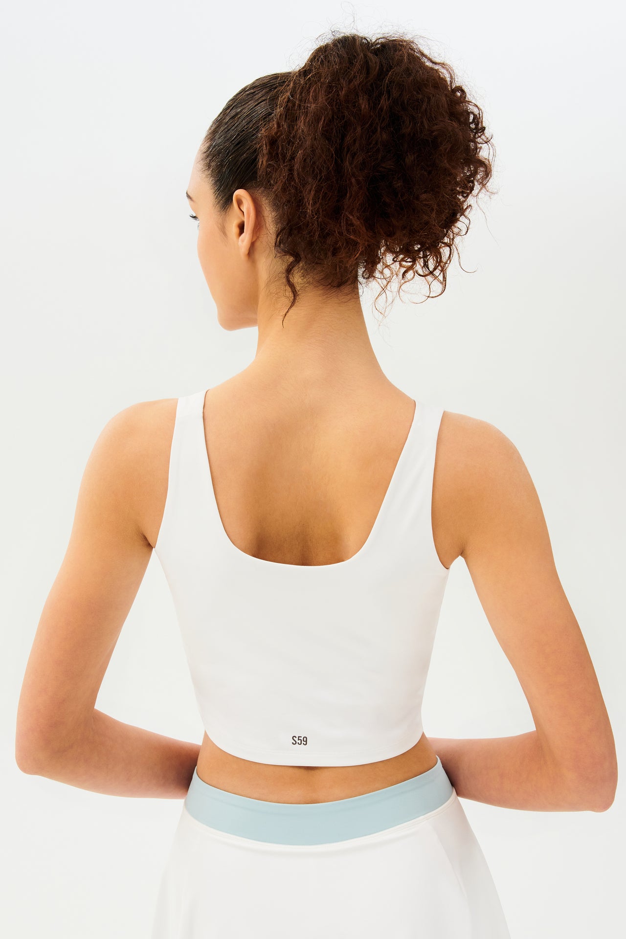 Back view of woman with dark curly hair in a ponytail wearing a cropped white tank bra with black S59 lettering and white skort with teal details on the waistband and white skort with tea waistband