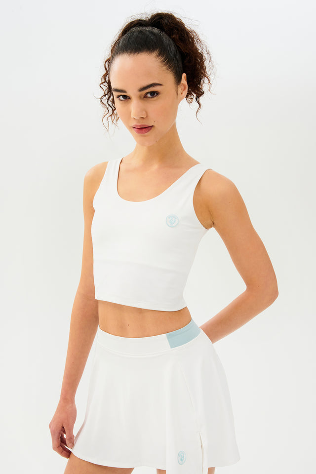 Front full view of woman with dark curly hair in a ponytail wearing a cropped white tank bra with white skort with teal details on the waistband and logo