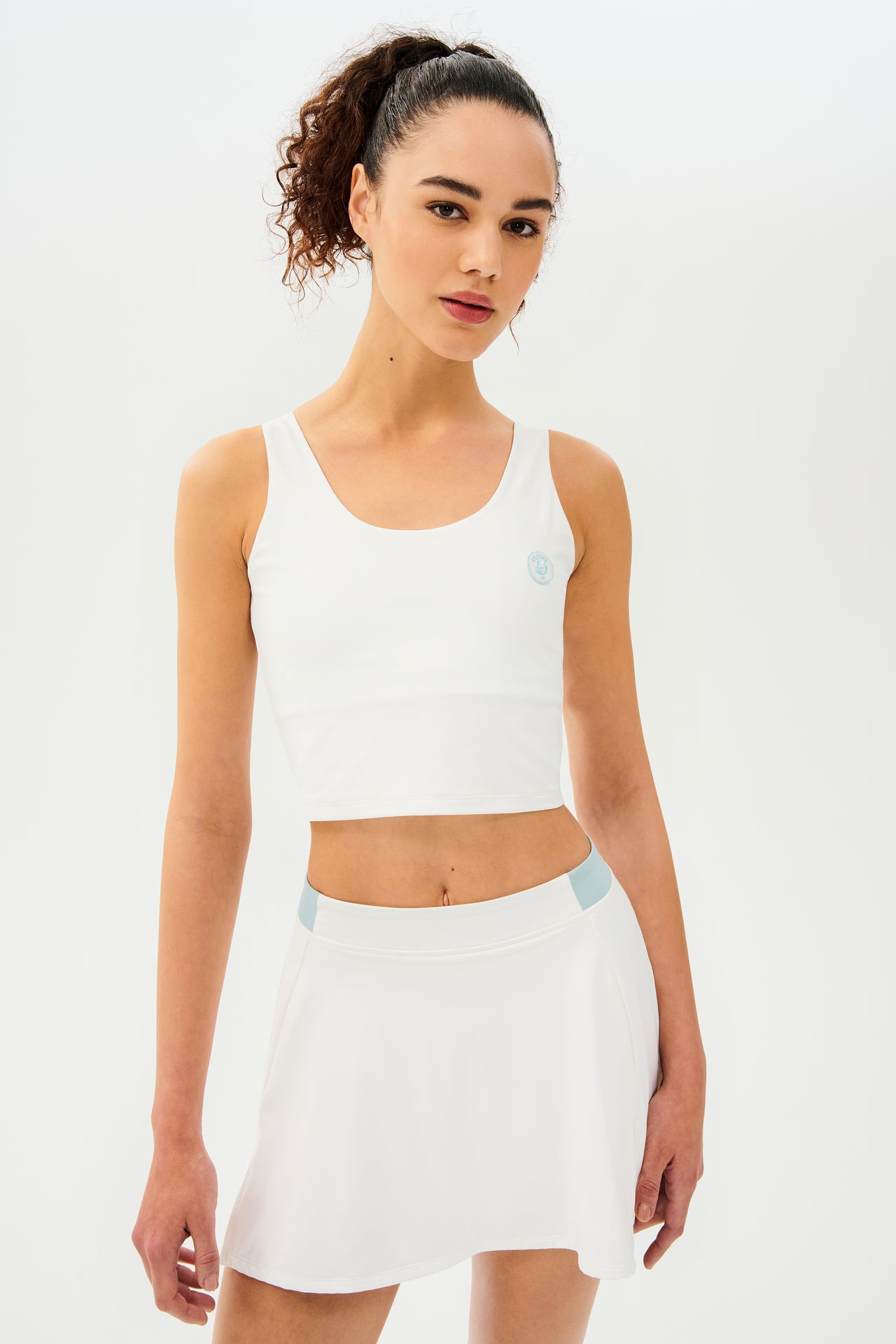 Front side view of woman with dark curly hair in a ponytail wearing a cropped white tank bra with white skort with teal details on the waistband and logo