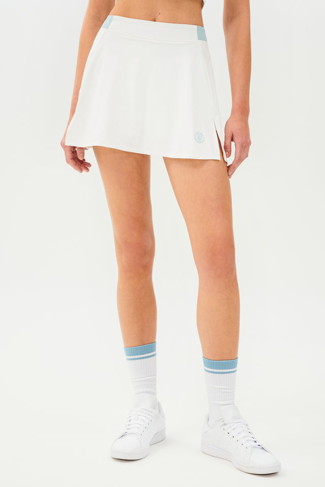 Front full view of white skort with teal details on the waistband and logo. Model is wearing white socks with teal stripe and all white shoes