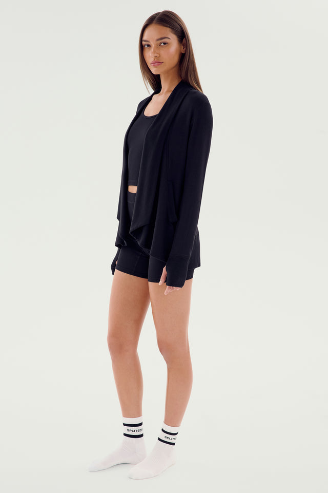 A person stands against a light background, wearing a black Celine Fleece Cardigan by SPLITS59 along with a crop top and shorts—an active wardrobe staple—paired with white socks and shoes.