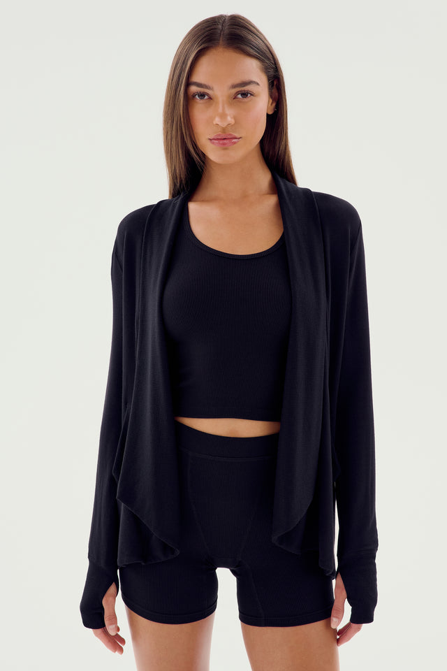 A woman stands against a plain background wearing an active wardrobe staple: the SPLITS59 Celine Fleece Cardigan in black, paired with a black crop top and black shorts, exuding effortless Los Angeles chic.