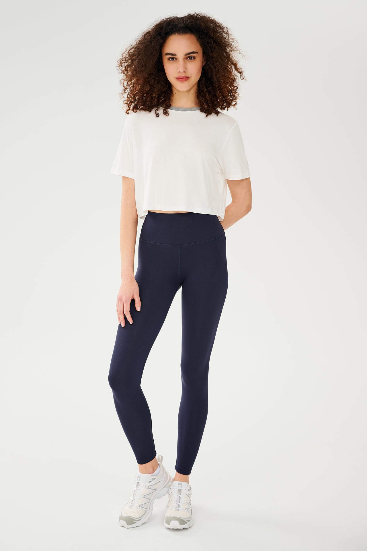 Full front view of girl wearing white cropped short sleeve t-shirt with thin light grey neck hem and dark blue leggings with white shoes