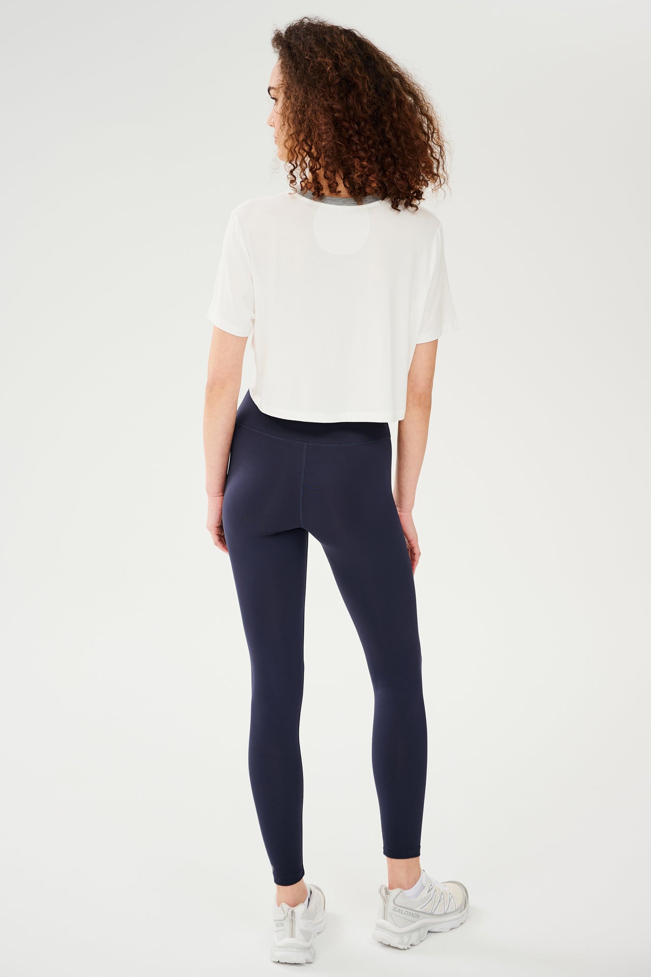 Full back view of girl wearing white cropped short sleeve t-shirt with thin light grey neck hem and dark blue leggings with white shoes