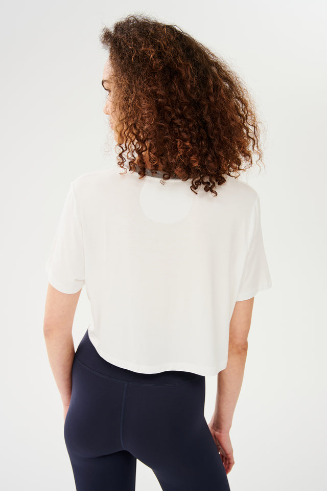 Back view of girl wearing white cropped short sleeve t-shirt with thin light grey neck hem and dark blue leggings 