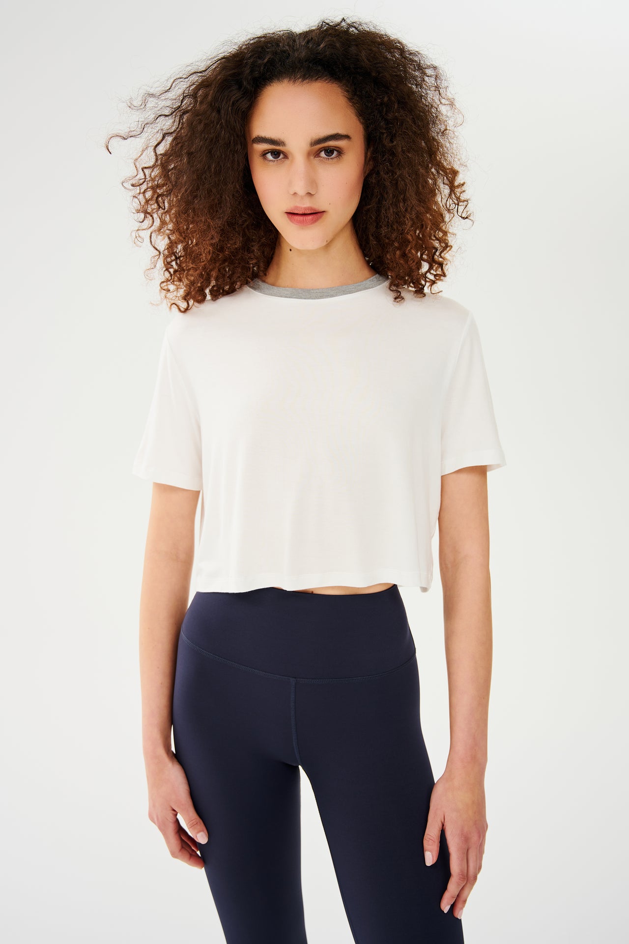 Front view of girl wearing white cropped short sleeve t-shirt with thin light grey neck hem and dark blue leggings 