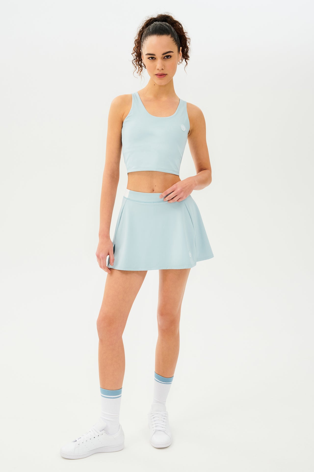 Front full view of woman with dark curly hair in a ponytail wearing a cropped teal tank bra with teal skort with white details on the waistband and logo. Model is wearing white socks with teal stripe and all white shoes