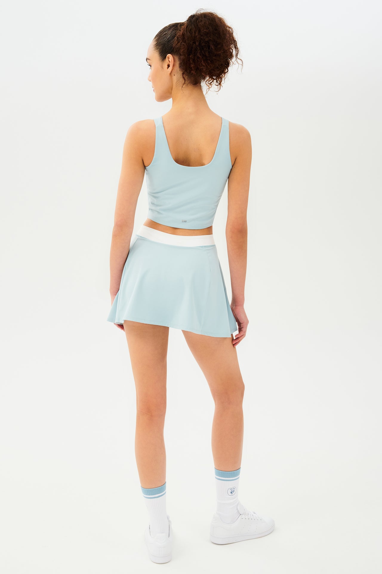 Back full view of woman with dark curly hair in a ponytail wearing a cropped teal tank bra with teal skort with white details on the waistband and logo. Model is wearing white socks with teal stripe and all white shoes
