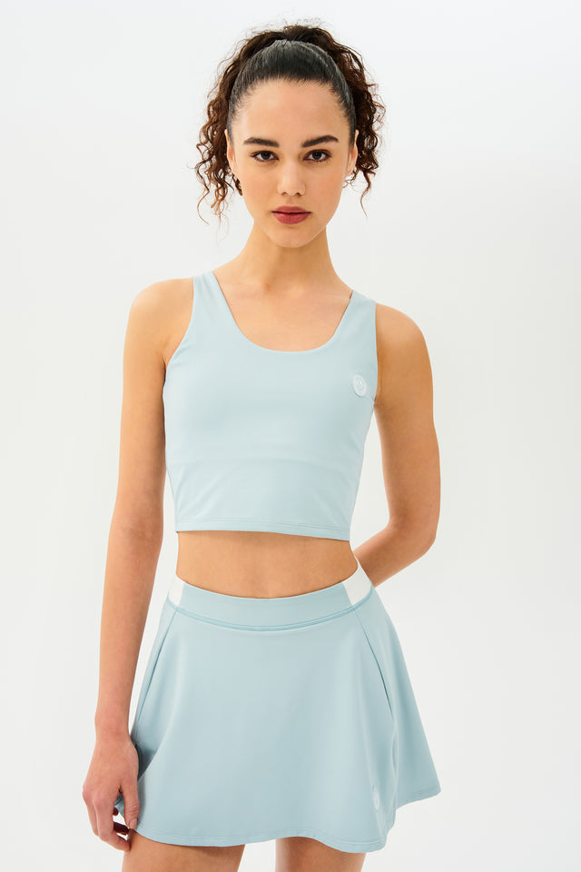 Front view of woman with dark curly hair in a ponytail wearing a cropped teal tank bra with teal skort with white details on the waistband and logo.