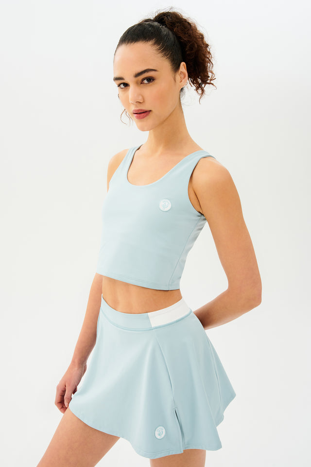 Front side view of woman with dark curly hair in a ponytail wearing a cropped teal tank bra with teal skort with white details on the waistband and logo.