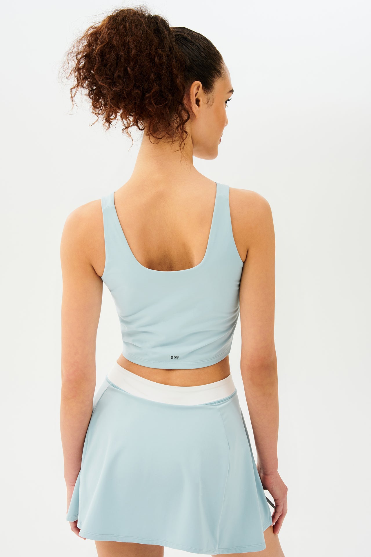 Back view of woman with dark curly hair in a ponytail wearing a cropped teal tank bra with teal skort with white strip on the waistband