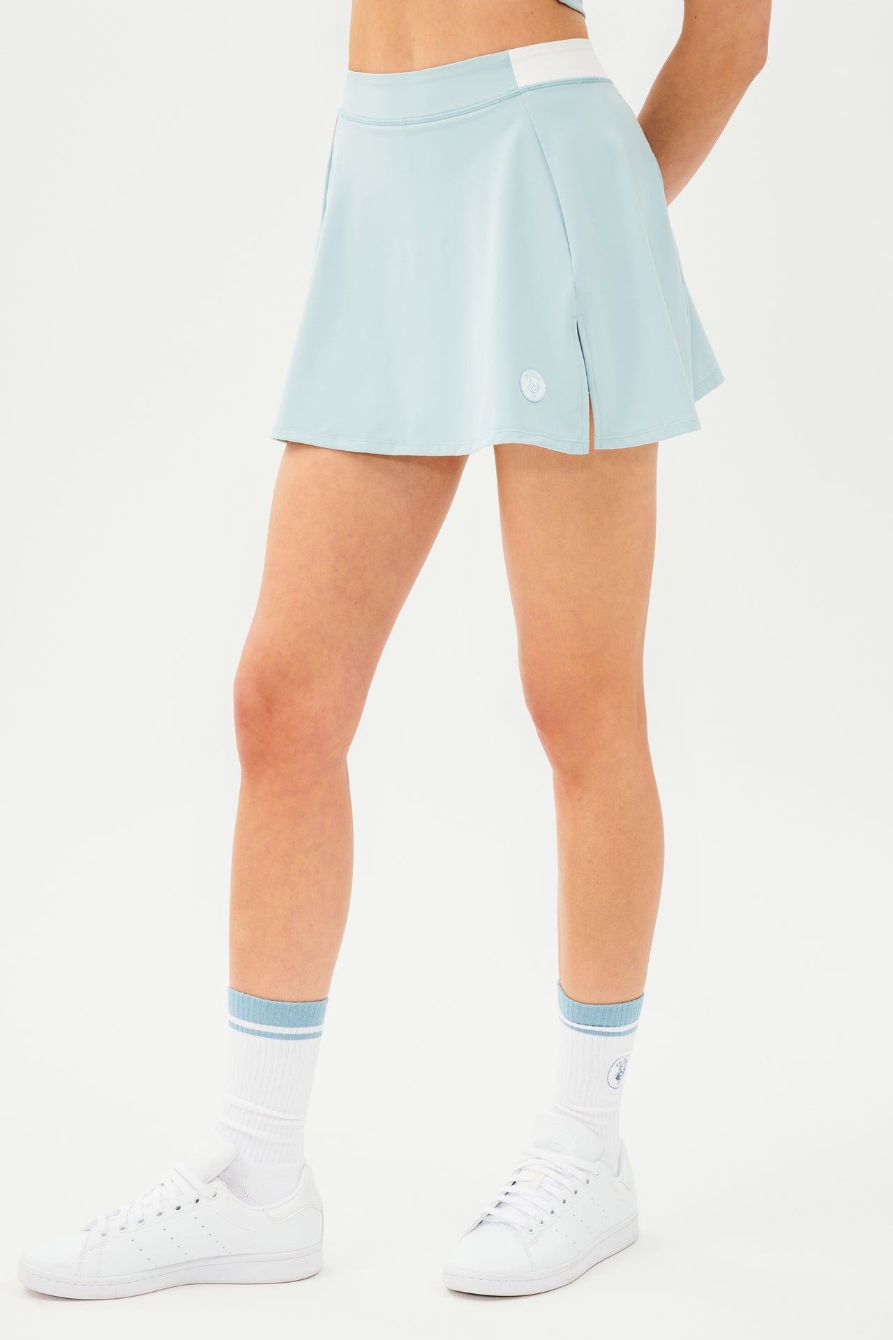 Front side view of teal skort with white details on the waistband. Model is wearing white socks with teal stripe and all white shoes