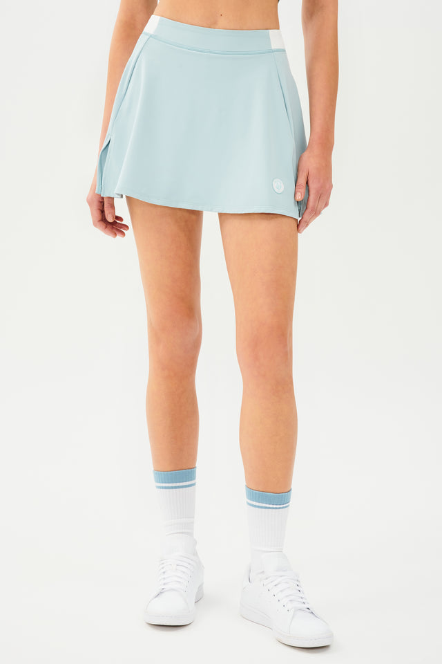 Front full view of teal skort with white  details on the waistband and logo. Model is wearing white socks with teal stripe and all white shoes