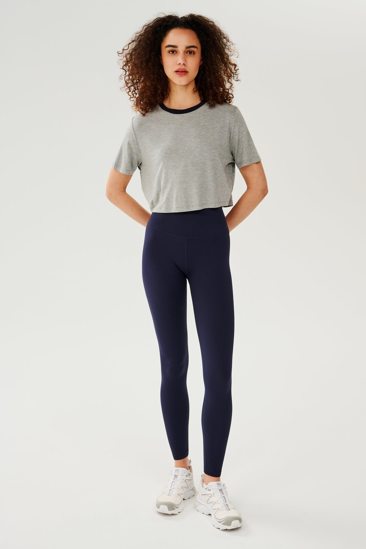 Full front view of girl wearing  light grey cropped short sleeve t-shirt with thin dark blue neck hem and dark blue leggings with white shoes