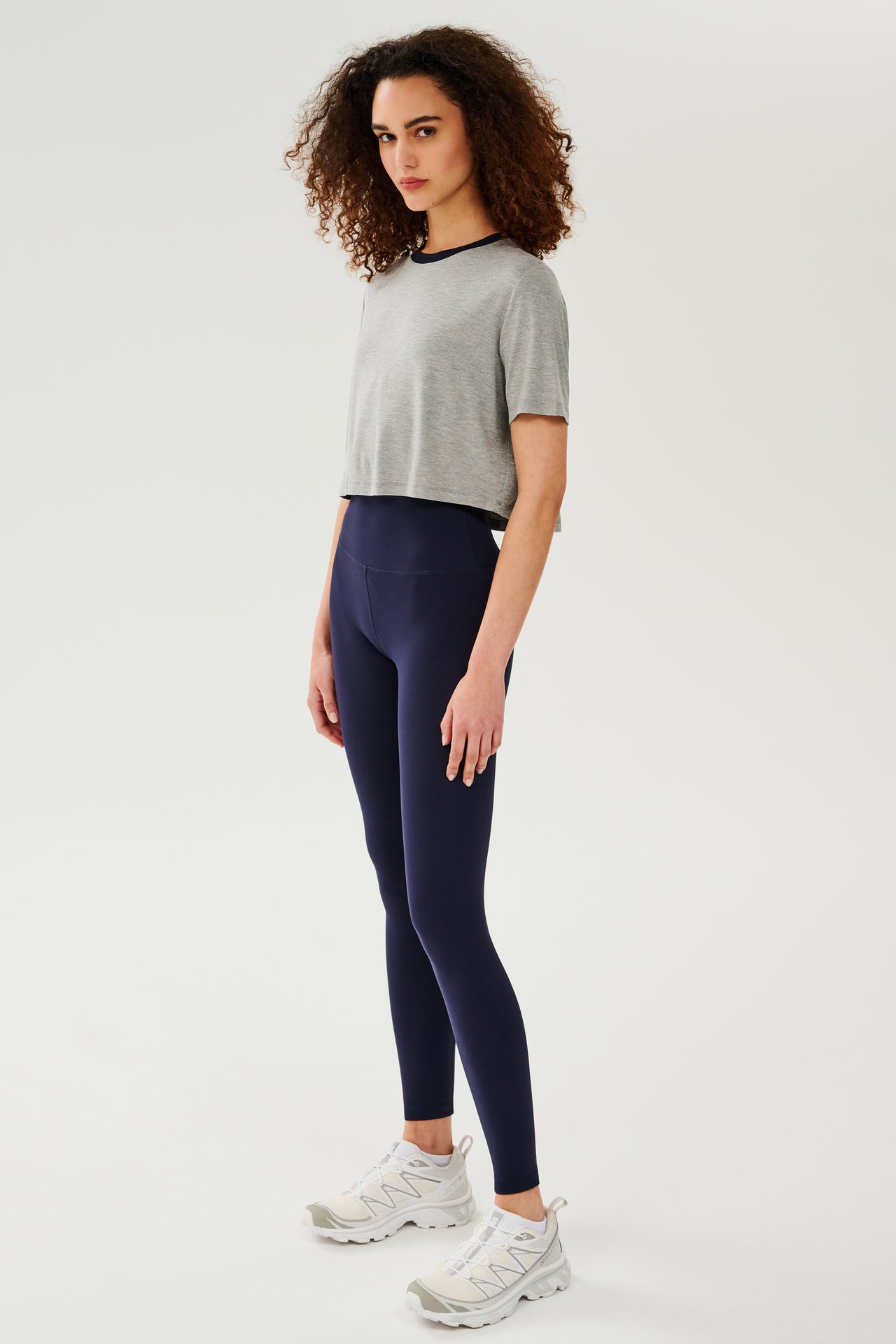 Full side view of girl wearing  light grey cropped short sleeve t-shirt with thin dark blue neck hem and dark blue leggings with white shoes