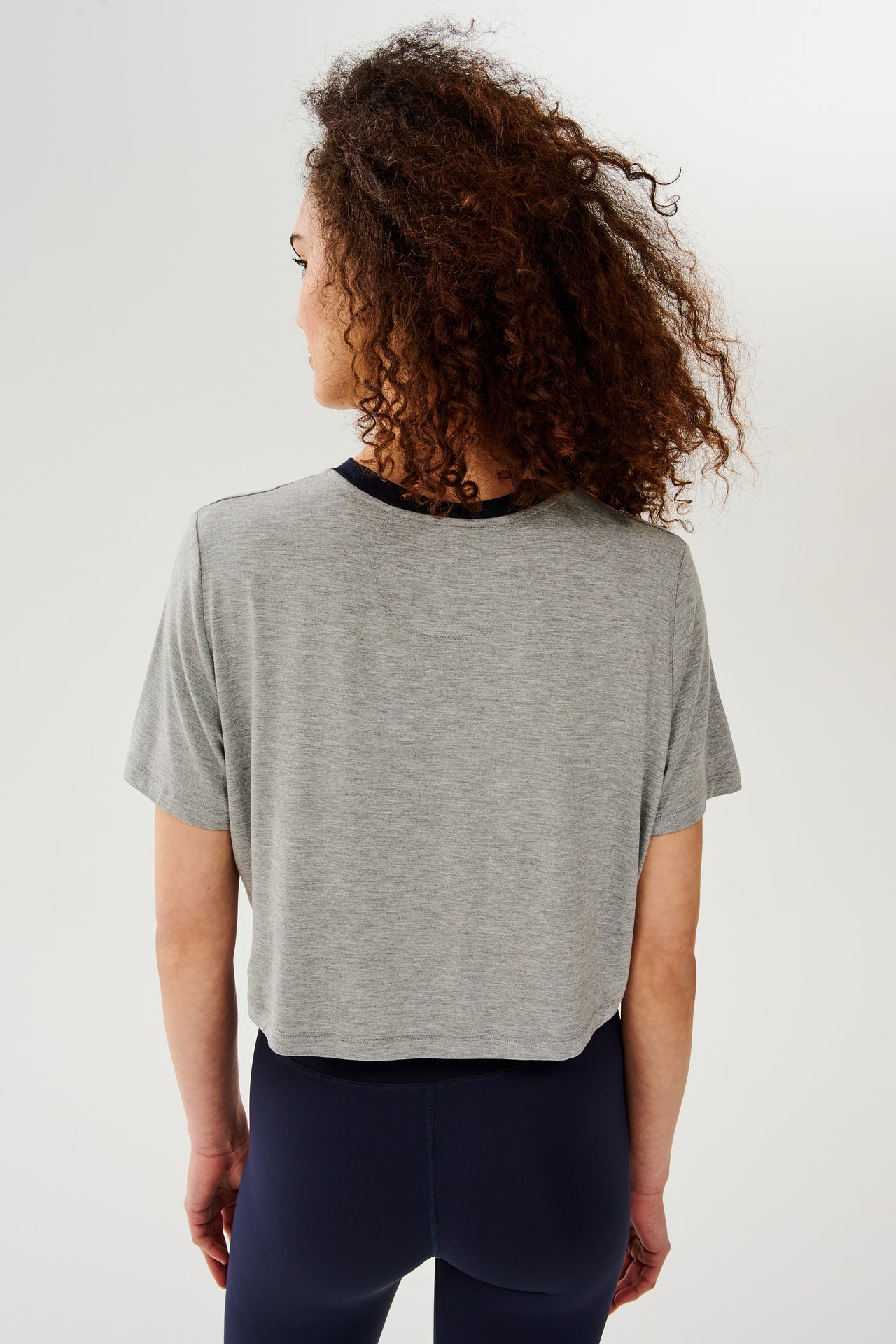 Back view of girl wearing  light grey cropped short sleeve t-shirt with thin dark blue neck hem and dark blue leggings 