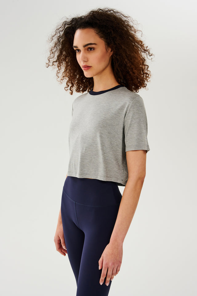 Side view of girl wearing  light grey cropped short sleeve t-shirt with thin dark blue neck hem and dark blue leggings 