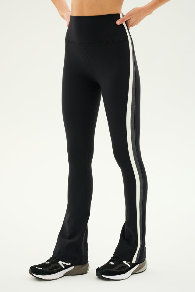 Person wearing SPLITS59's Raquel High Waist Flared Legging in black, white, and heather grey with a white side stripe and black athletic shoes, crafted from 4-way stretch supplex fabric, standing against a plain background.