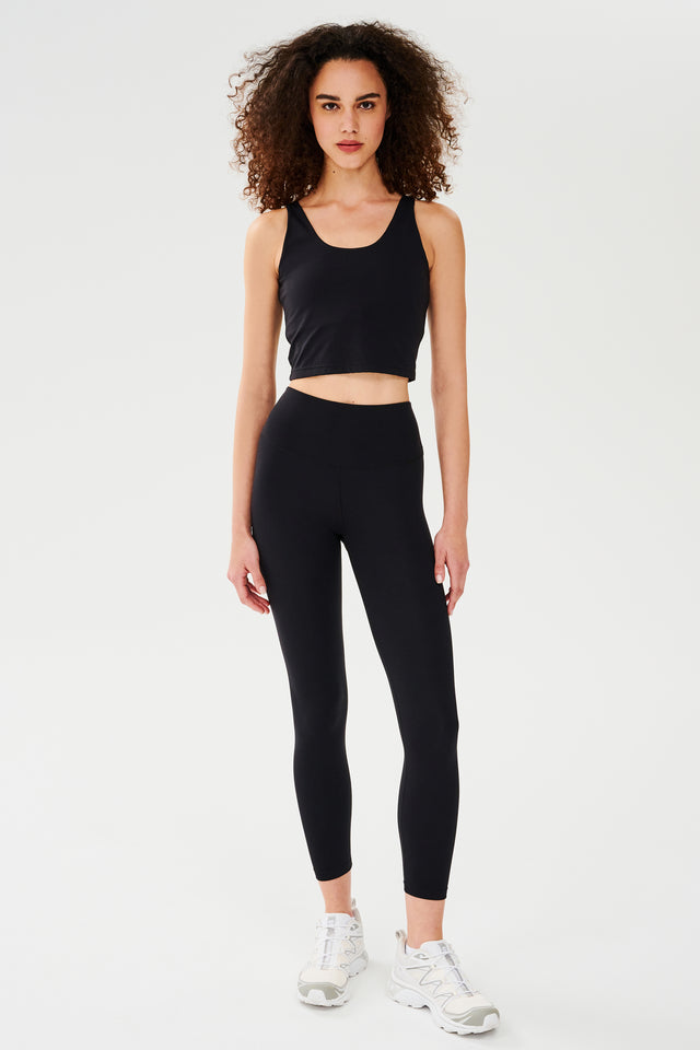 Front full view of woman with curly dark brown hair wearing a black bra crop tank with black leggings and white shoes 