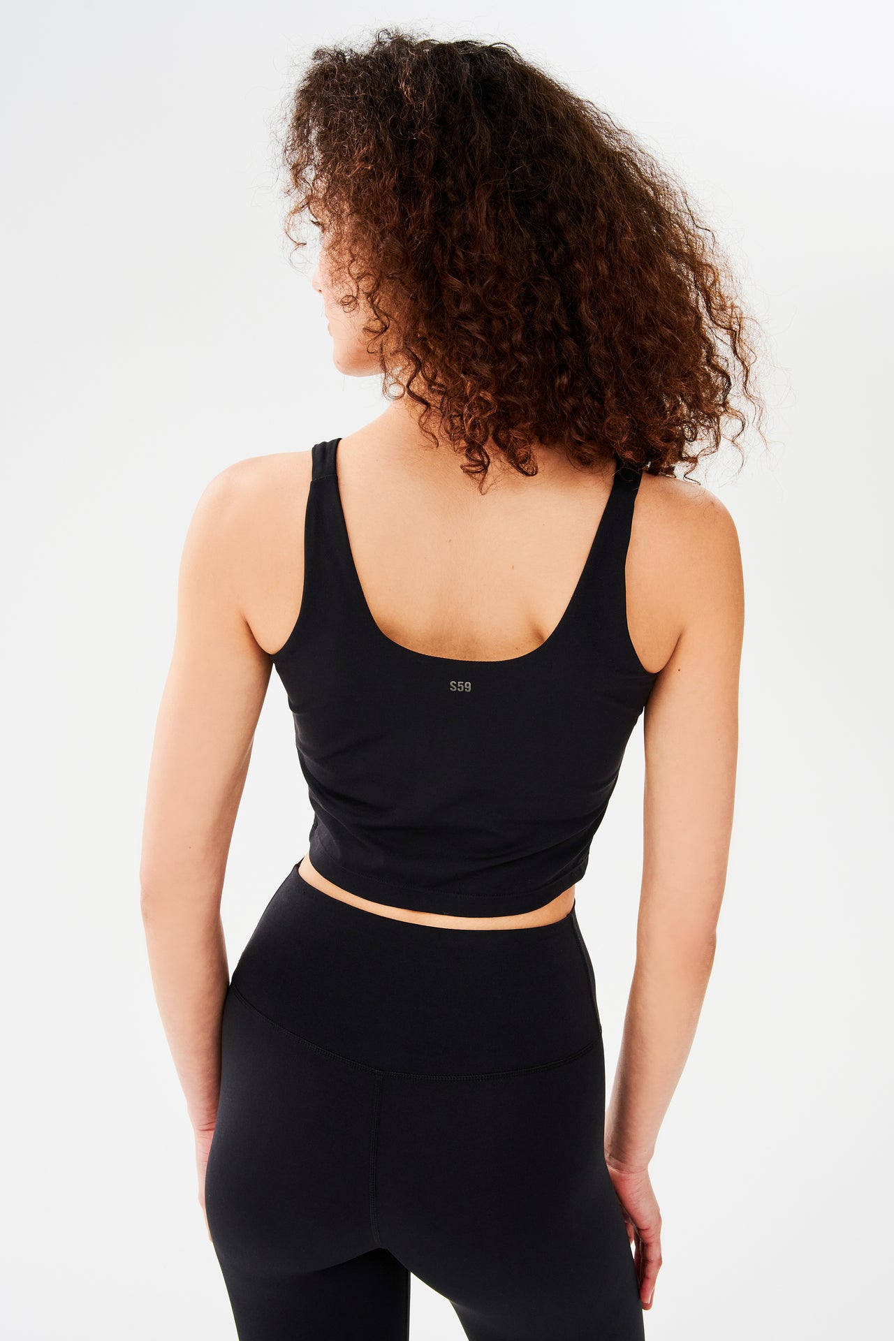 Back view of woman with curly dark brown hair wearing a black bra crop tank with  white S59 logo and black leggings