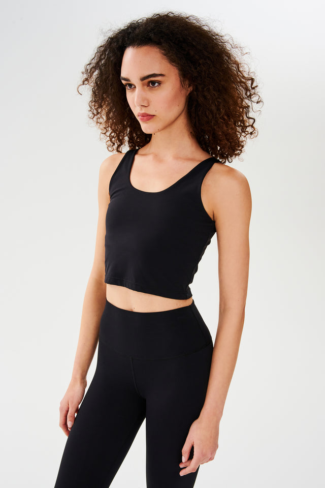 Front side view of woman with curly dark brown hair wearing a black bra crop tank with black leggings 